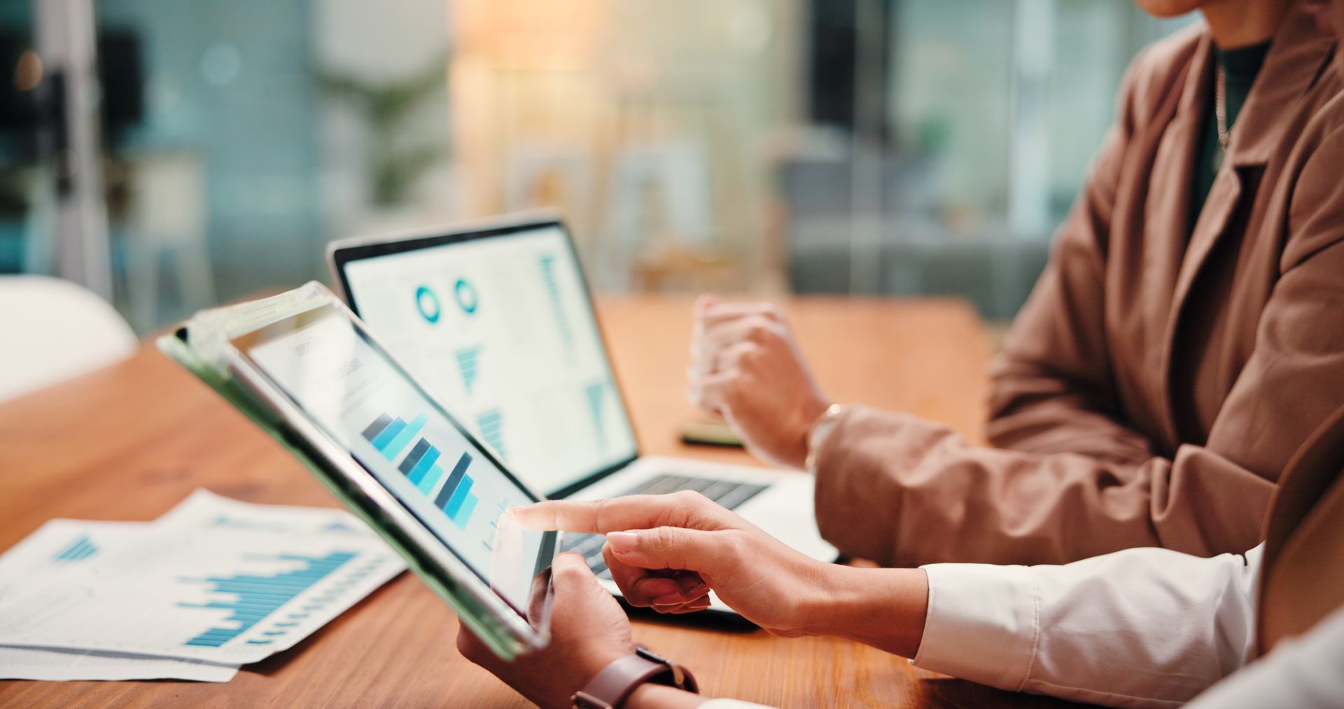 Two people analyze charts on a tablet and laptop at a wooden table in an office Two people analyze charts on a tablet and laptop at a wooden table in an office