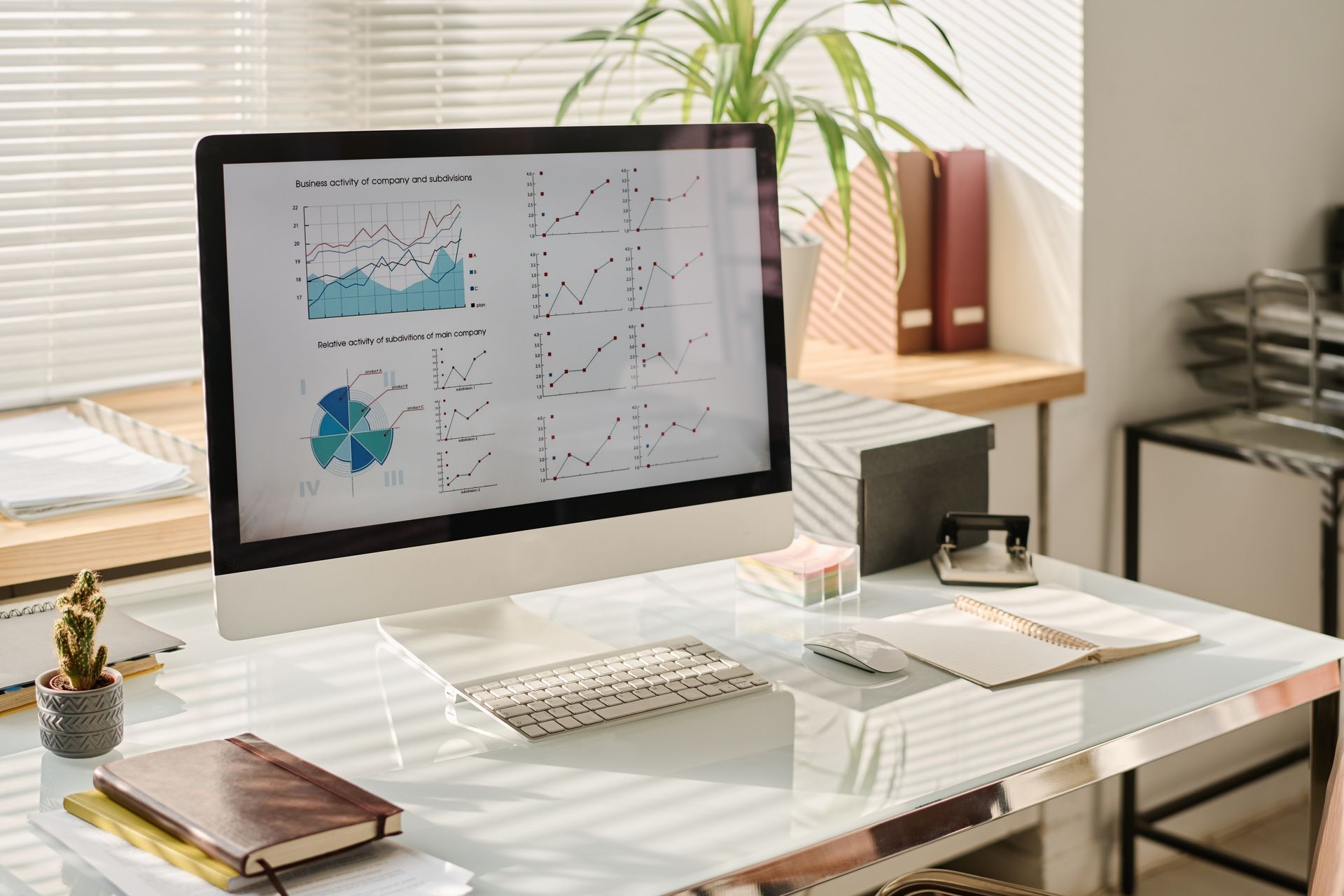Desktop computer showing charts, on a modern desk with office supplies and a plant, lit by sunlight Desktop computer showing charts, on a modern desk with office supplies and a plant, lit by sunlight