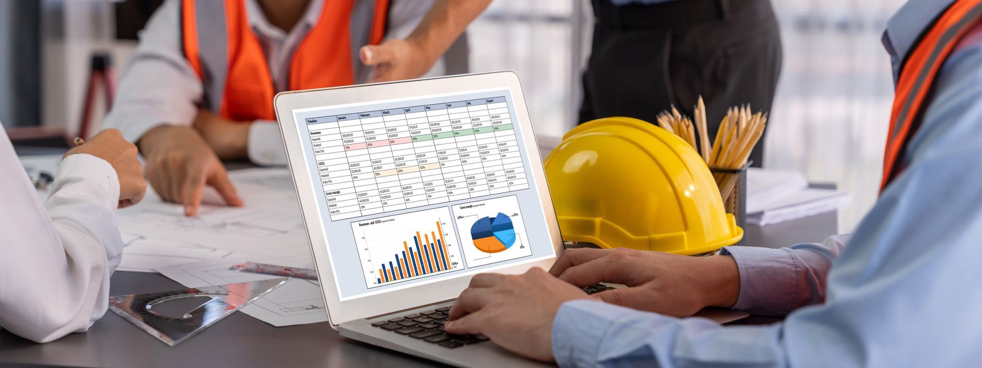 Construction workers review blueprints and data on a laptop, a hardhat and pencils on the table Construction workers review blueprints and data on a laptop, a hardhat and pencils on the table