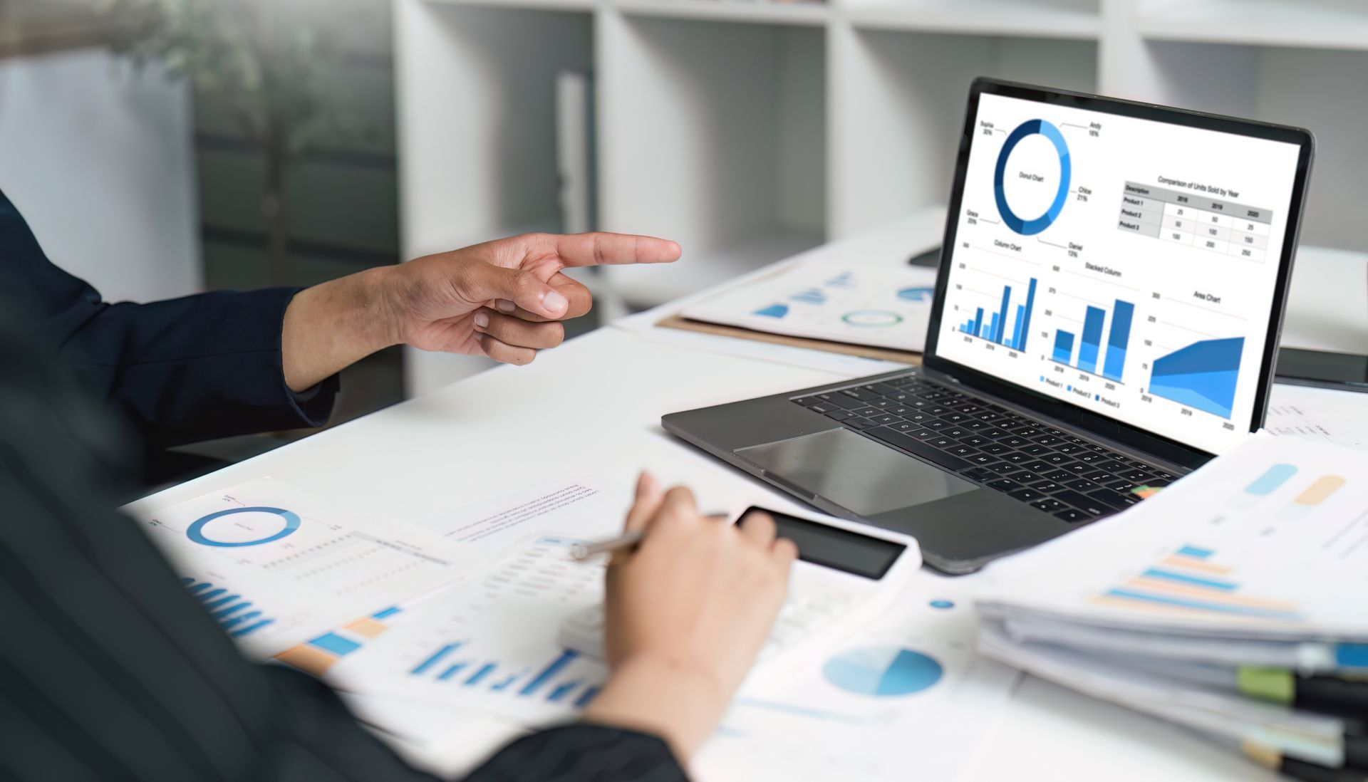 Person points at a laptop displaying financial data, using a calculator amid charts and documents Person points at a laptop displaying financial data, using a calculator amid charts and documents