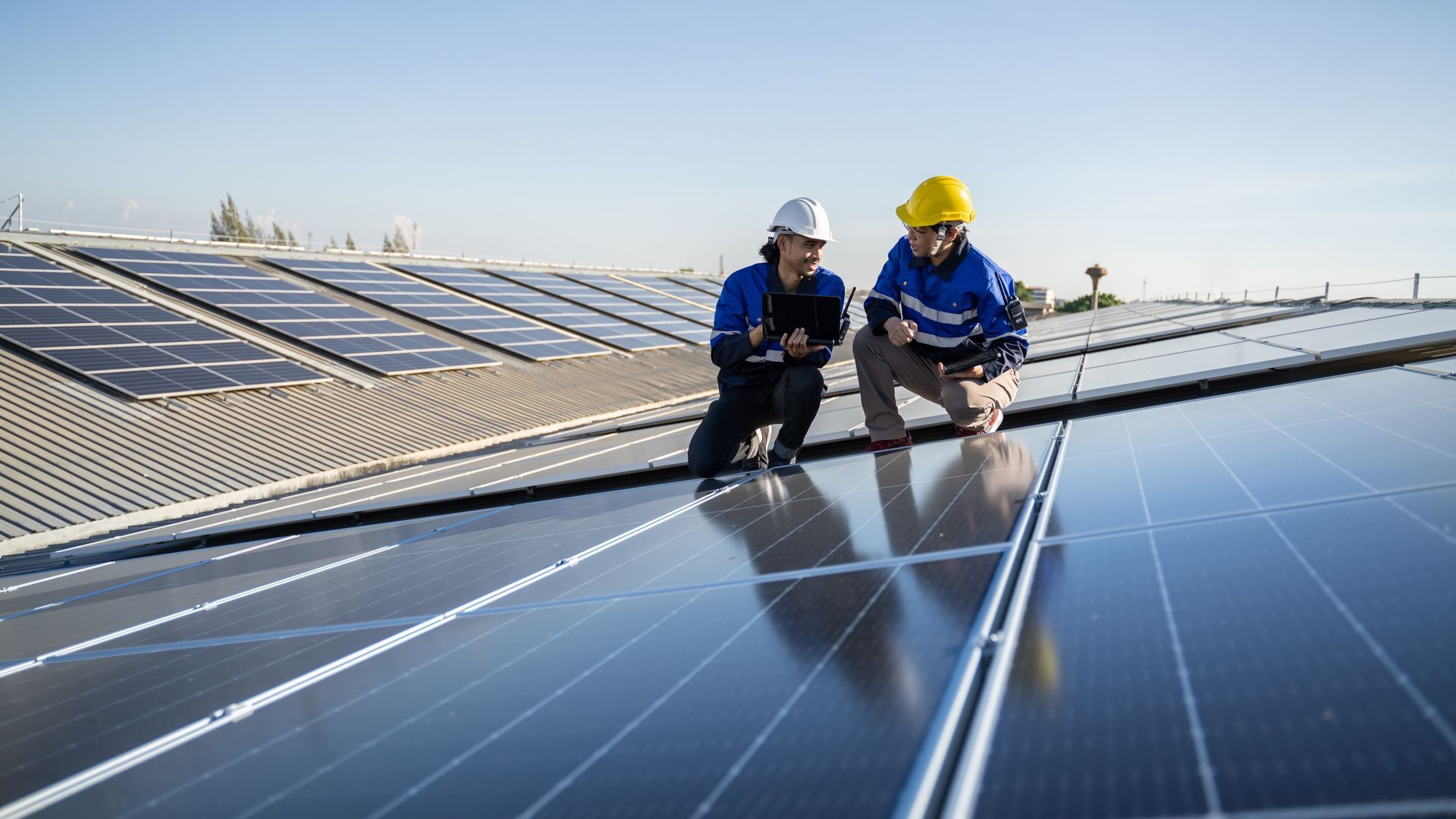 Two workers, one with a yellow hard hat and another with a white hard hat, inspecting solar panels on a rooftop Two workers, one with a yellow hard hat and another with a white hard hat, inspecting solar panels on a rooftop