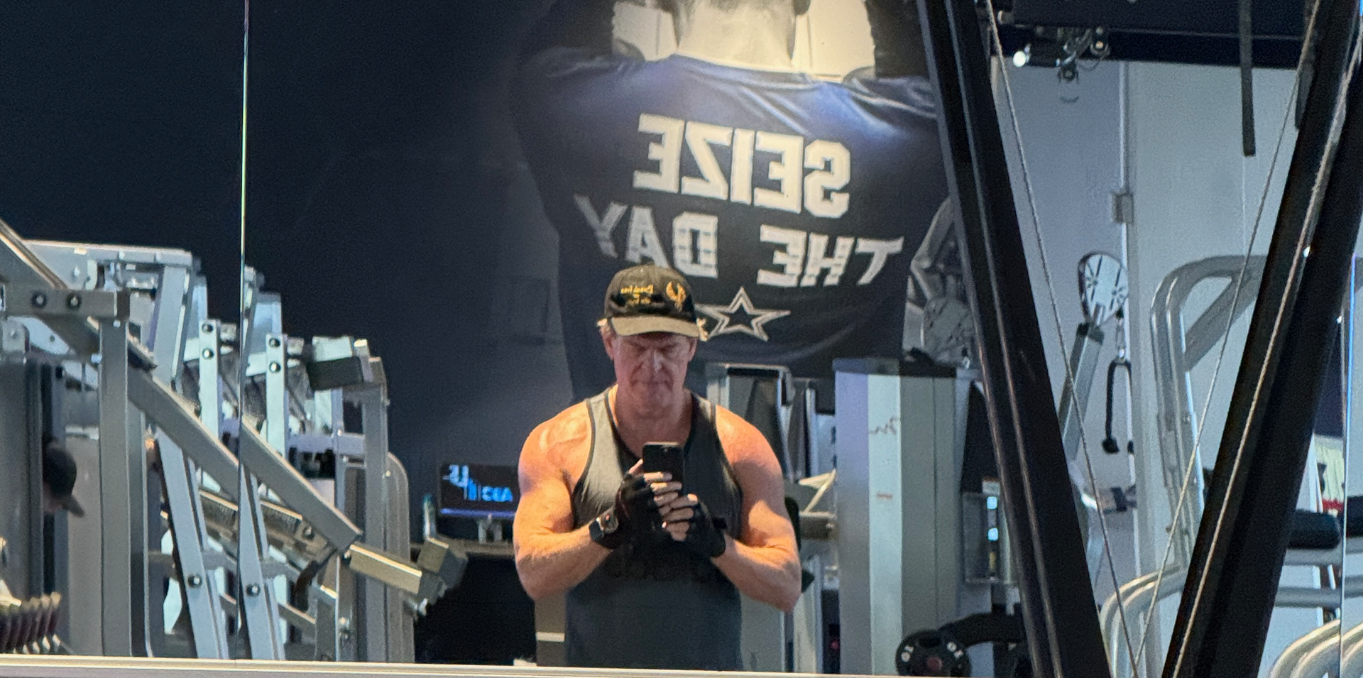 Man in gym wearing a hat and gloves, flexing his arms. 