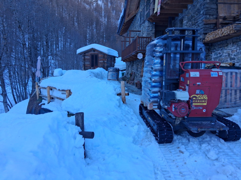 Un carrello elevatore cingolato rosso, carico di provviste, è parcheggiato davanti a un rustico edificio in pietra in un paesaggio boschivo innevato.