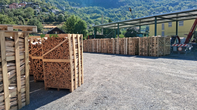 Casse di legno impilate e piene di legna da ardere, disposte in file su un piazzale ghiaioso sotto una tettoia vicino a una collina.