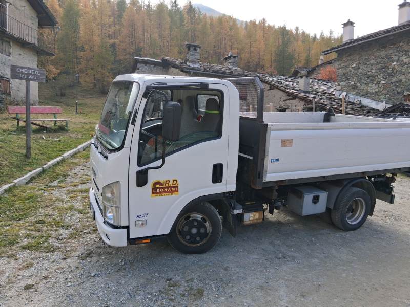 Un camion ribaltabile bianco parcheggiato su un sentiero di ghiaia di fronte a rustici edifici in pietra e a una collina boscosa.