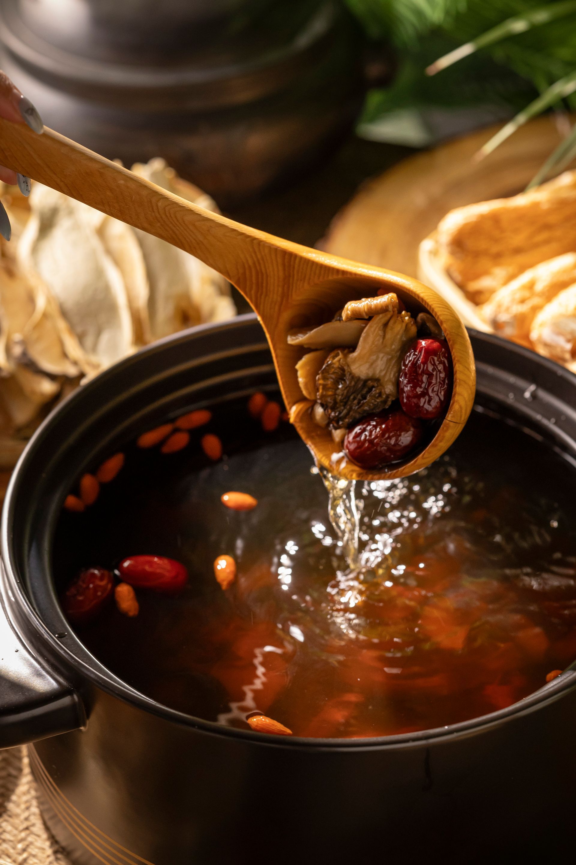 Wooden spoon lifting herbs from a black pot of simmering red broth with mushrooms