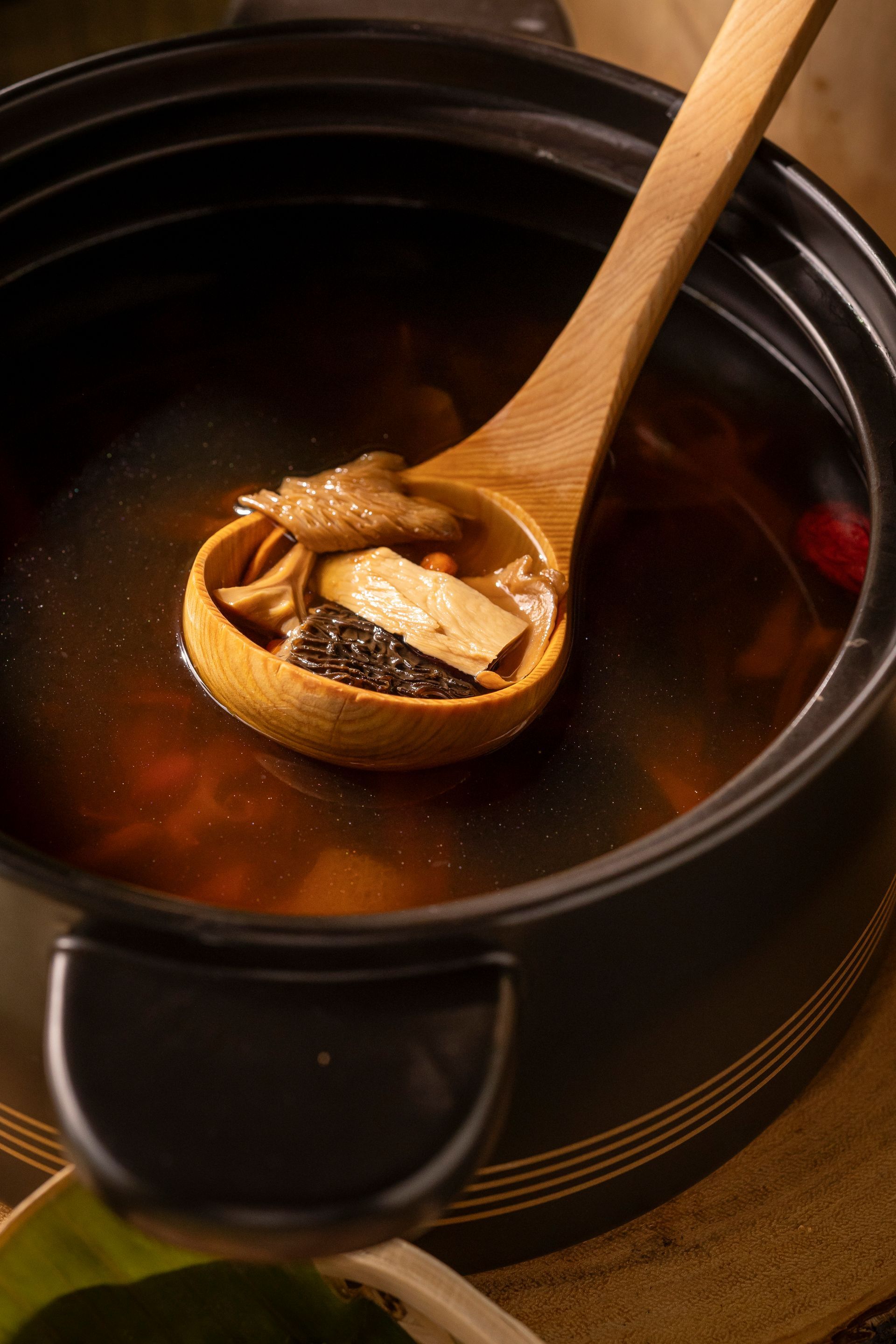 Wooden spoon lifting tofu from a dark ceramic pot of simmering broth