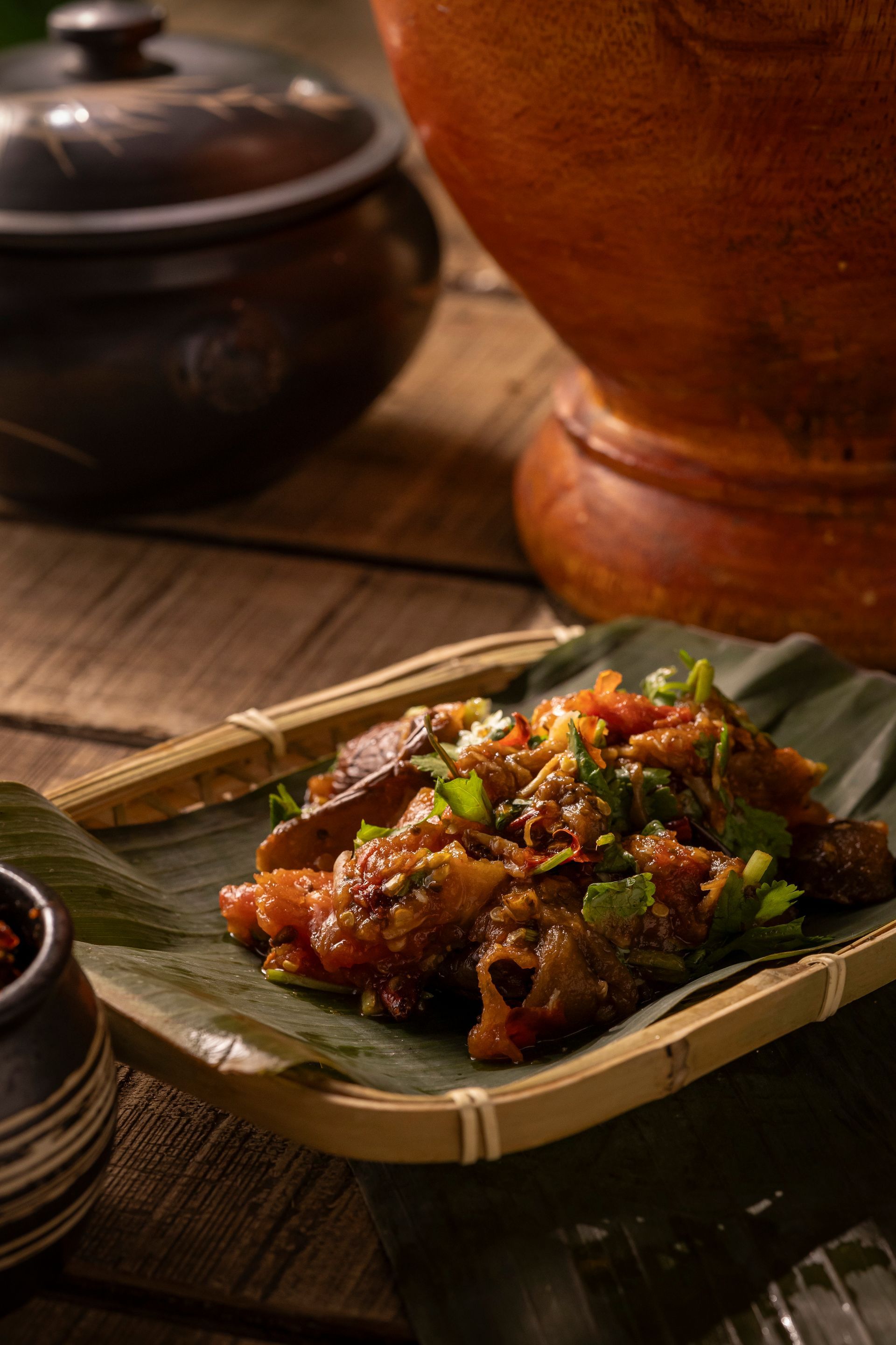 Grilled meat on a banana leaf tray with chili sauce, on a wooden table with pots in the background