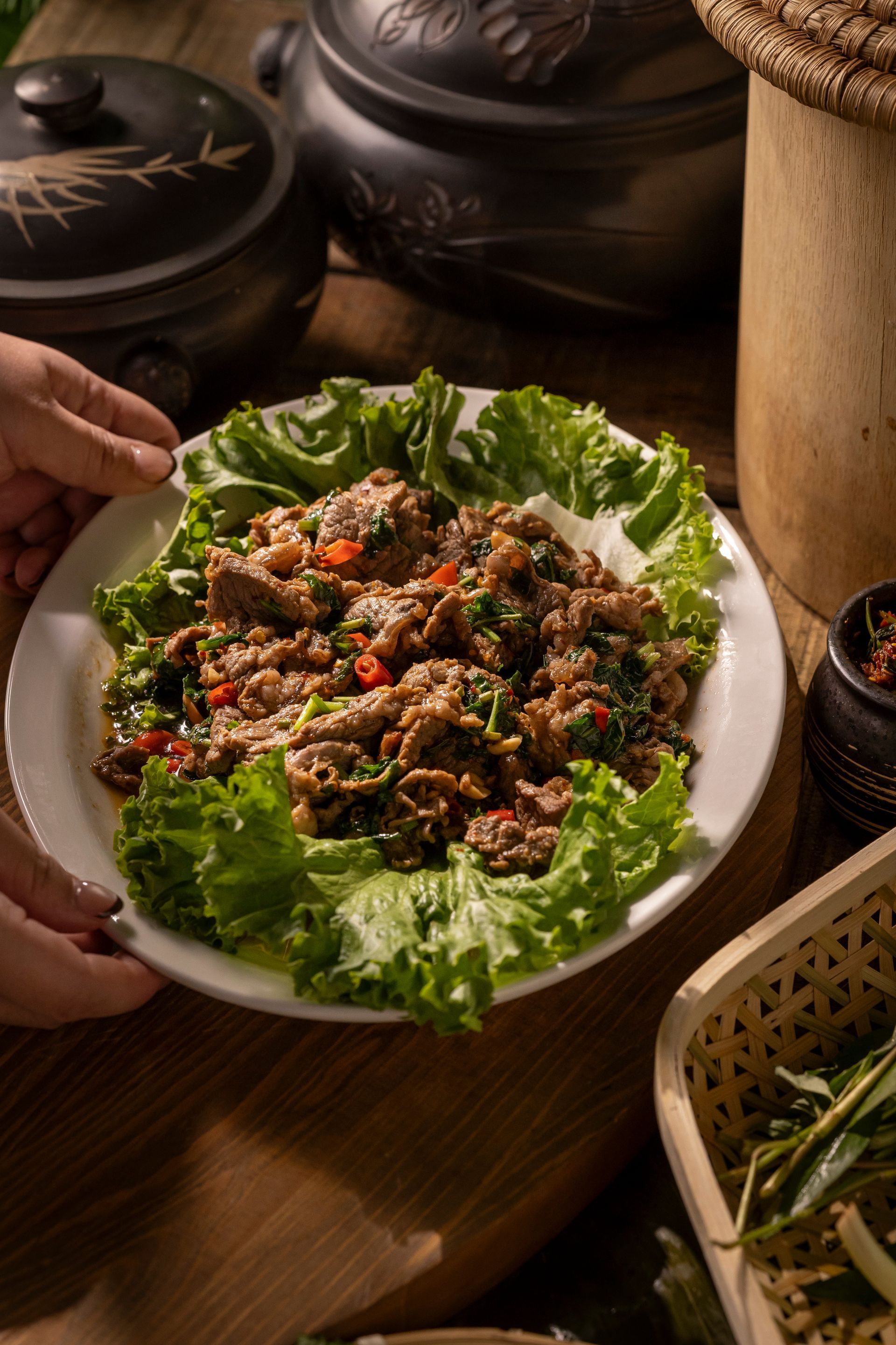 Hand-held plate of leafy salad topped with minced meat on a wooden table, with bowls and herbs nearby