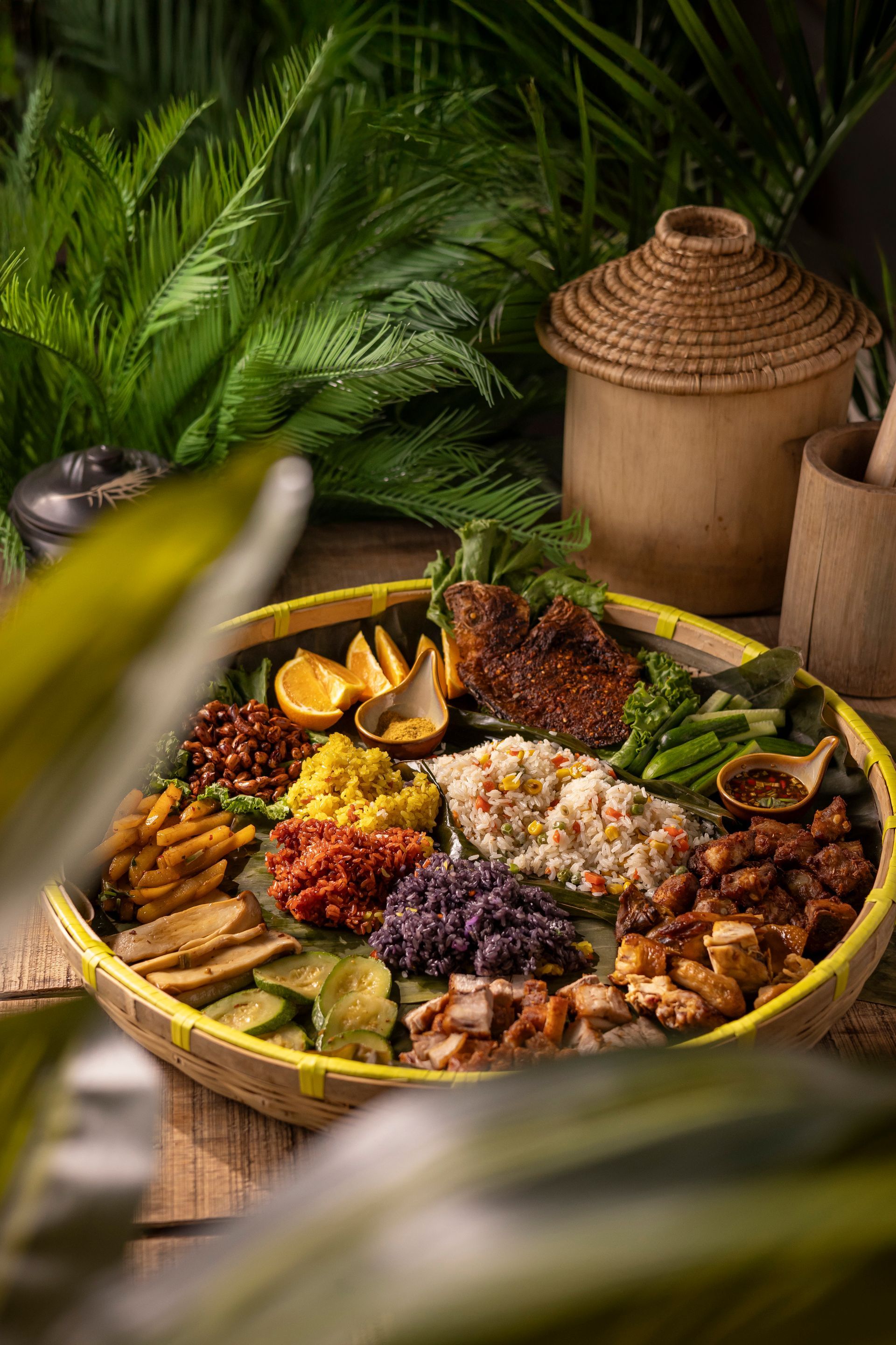 Colorful assortment of tropical fruits and dishes arranged on a large woven tray amid lush green leaves