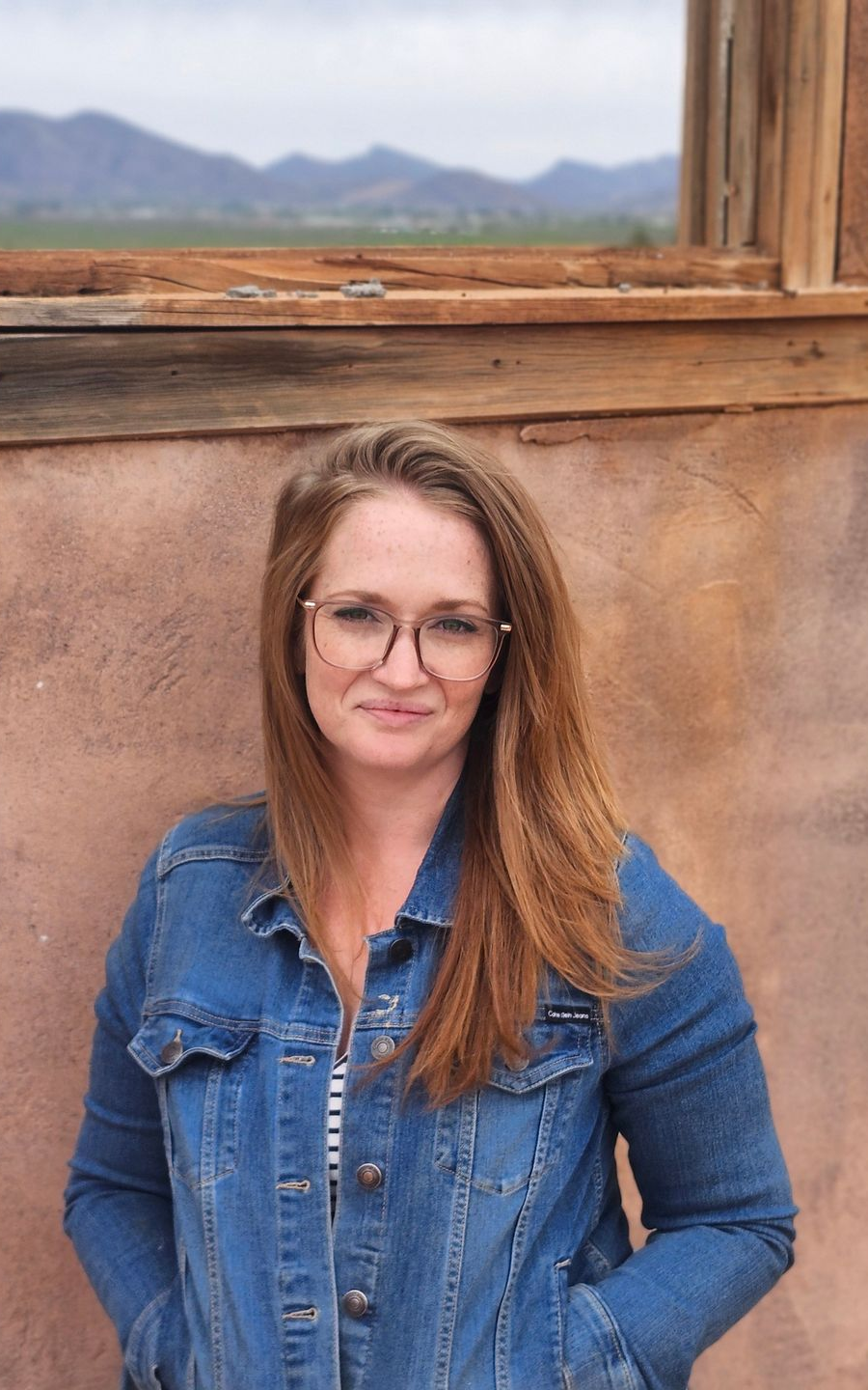 Woman in a denim shirt standing by a wooden window with mountains in the background