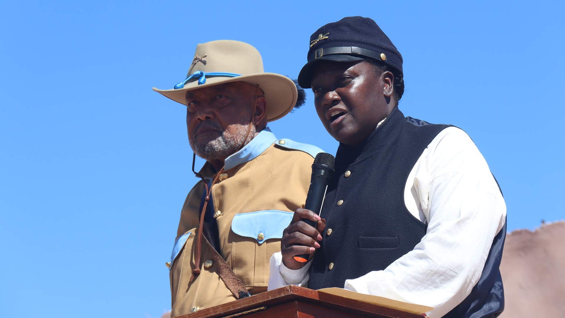 Two men in historical uniforms speaking at a podium outdoors.