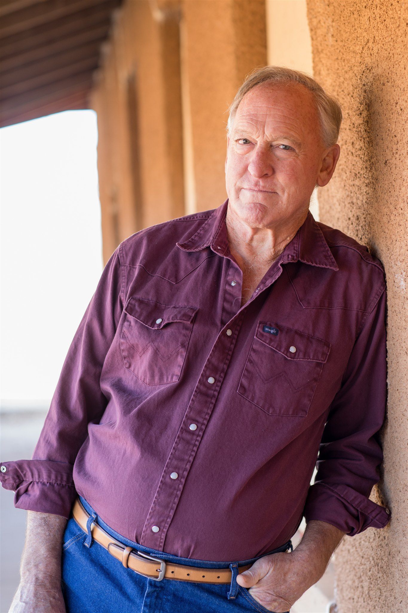 Man with gray hair and wrinkled face, looking at the camera. He is in front of a white wall.