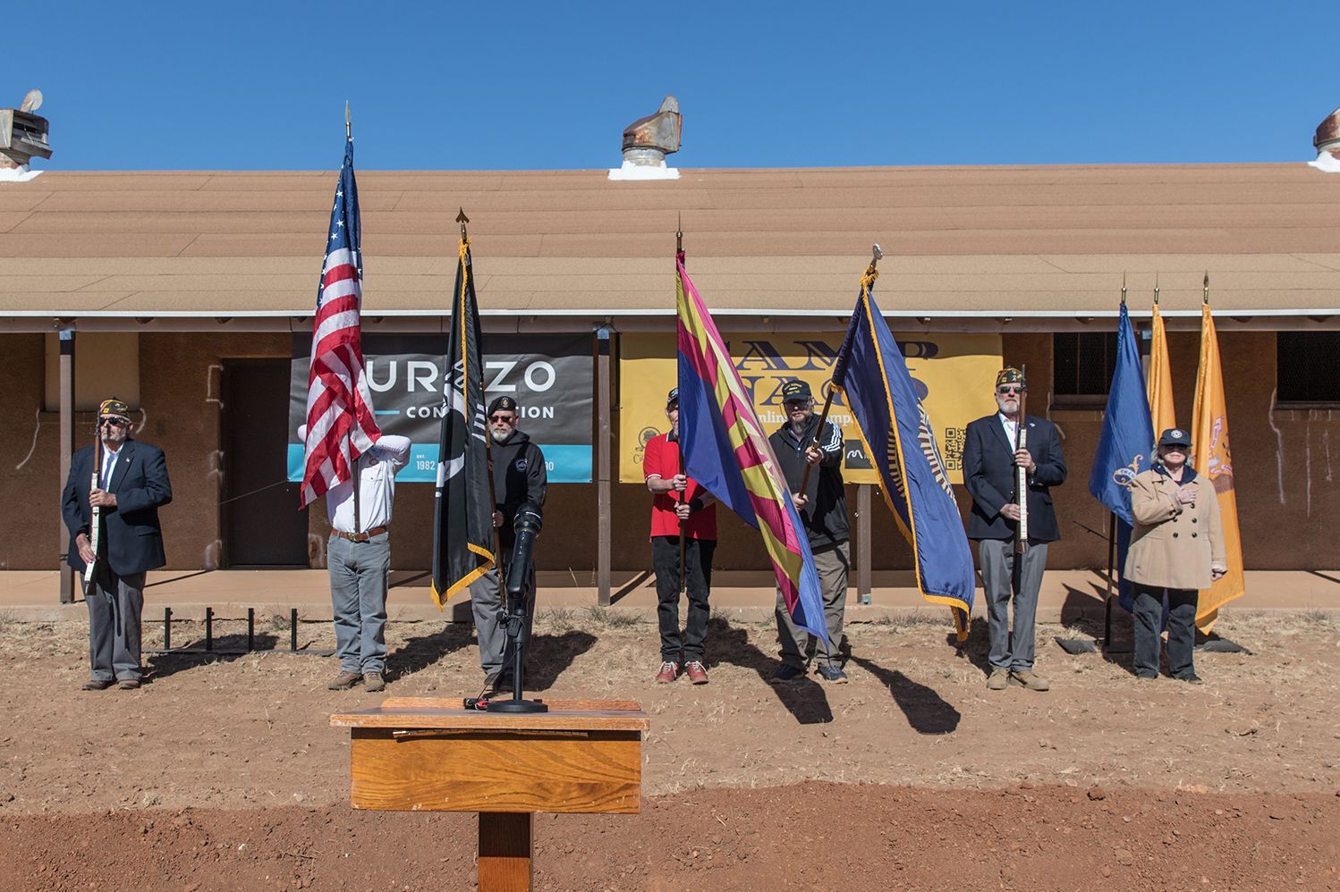 Group of people in uniform hold flags outdoors, standing in front of a building on a sunny day.