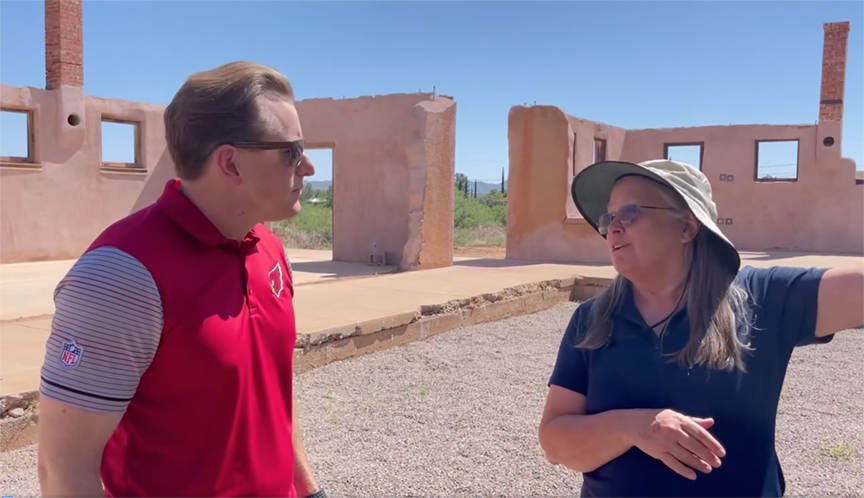 Man in red shirt and woman in sun hat conversing outside historic ruins on a sunny day.