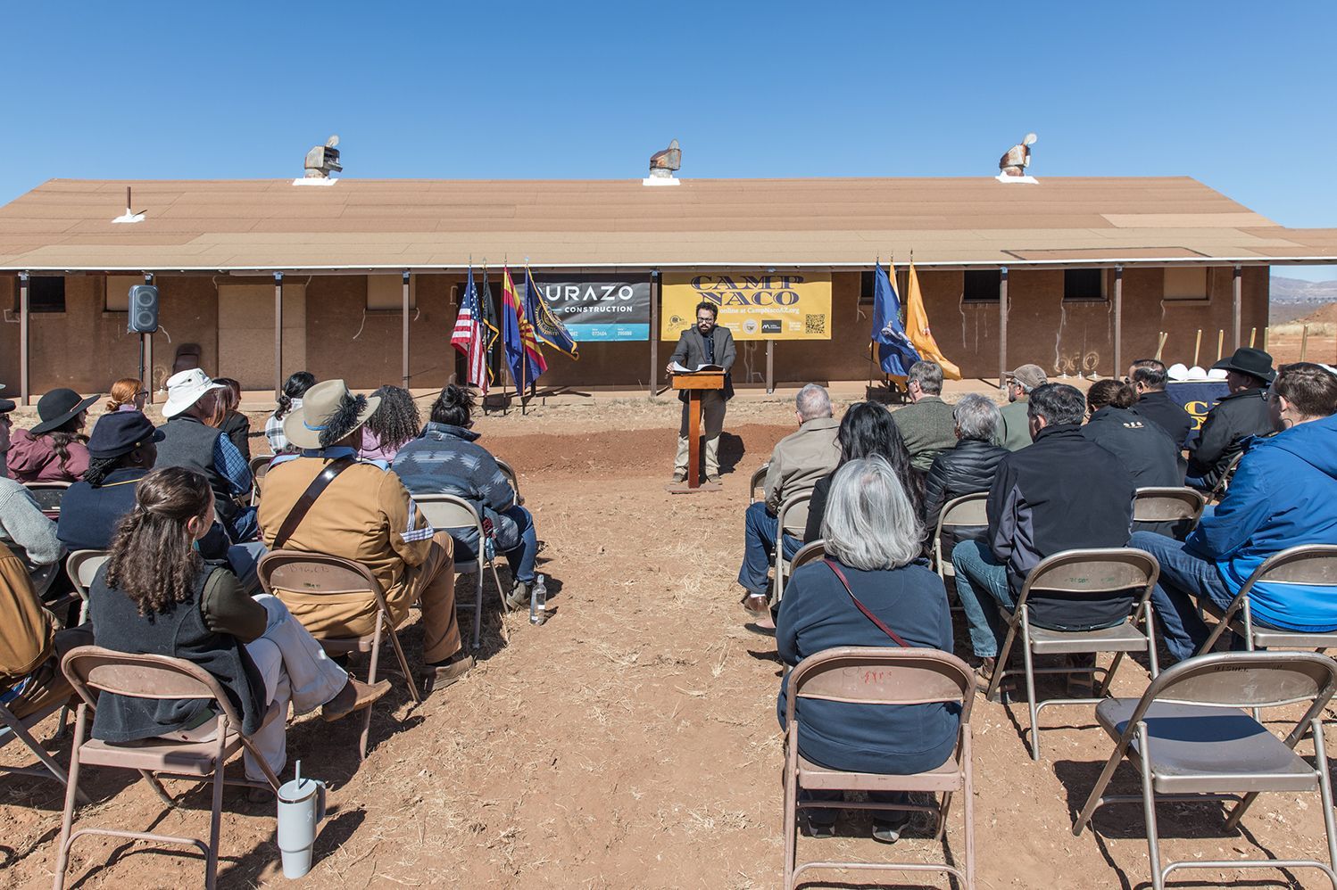 Man speaking at podium in front of a crowd seated on chairs outdoors at a building.