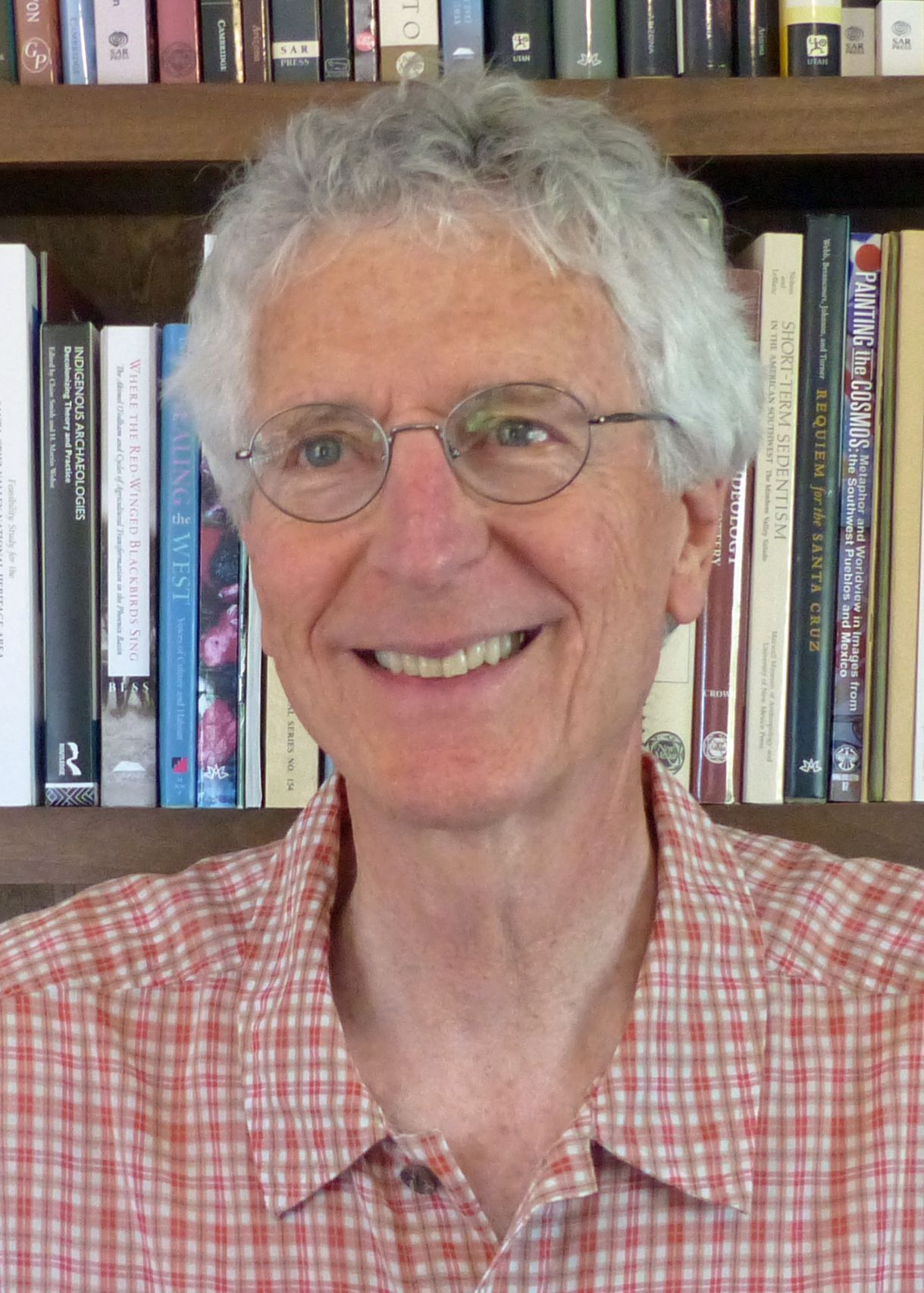 Man with gray hair and glasses smiling in front of a bookshelf. He wears a red and white plaid shirt.