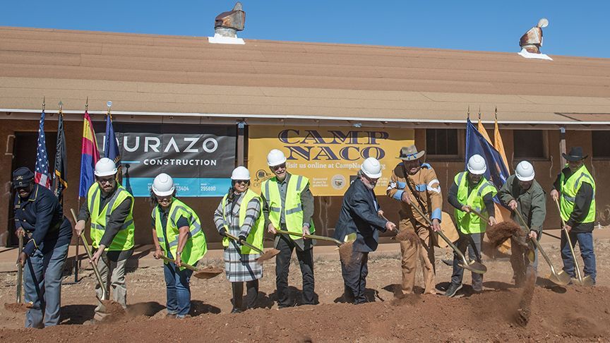 Group of people in hard hats and vests breaking ground with shovels in front of a building.