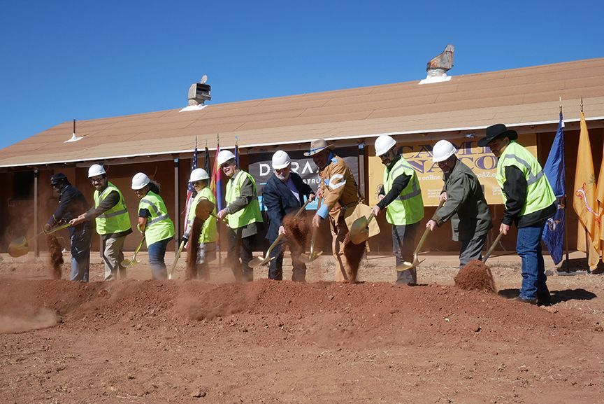 People in hard hats and vests shoveling dirt at a groundbreaking ceremony, brown building and flags in background.