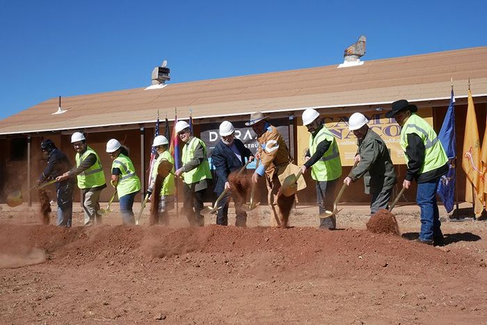 People in hard hats and vests shoveling dirt at a groundbreaking ceremony, brown building and flags in background.
