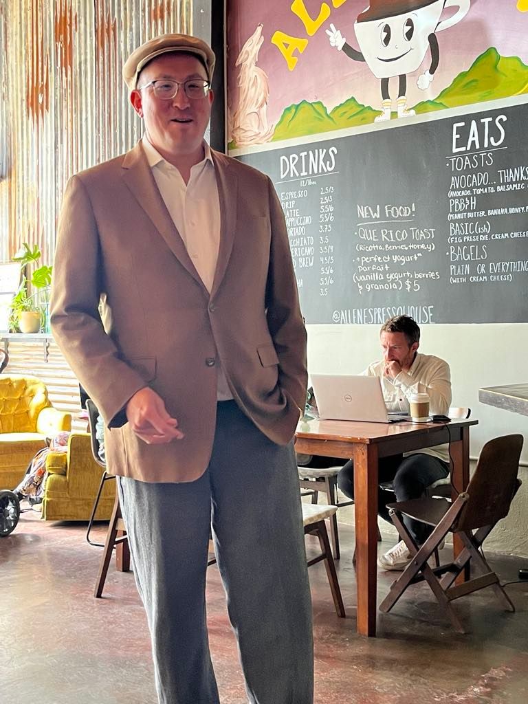 Man in a blazer and cap stands in a coffee shop, another person works at a table in the background.