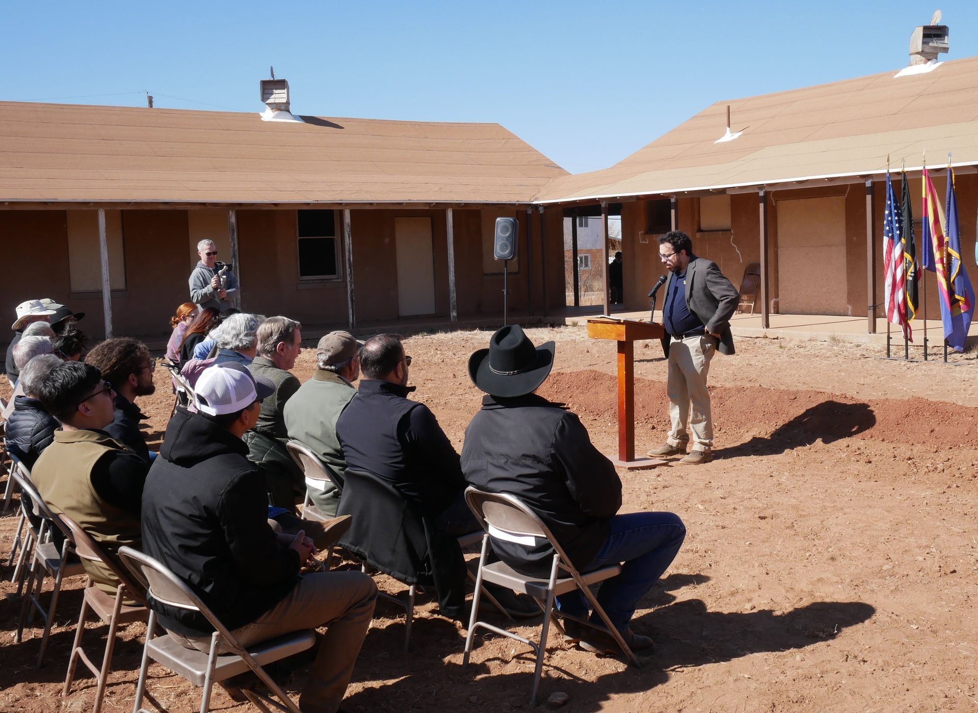 A man speaks at a podium to a seated audience outdoors, flags behind him. Buildings in background.