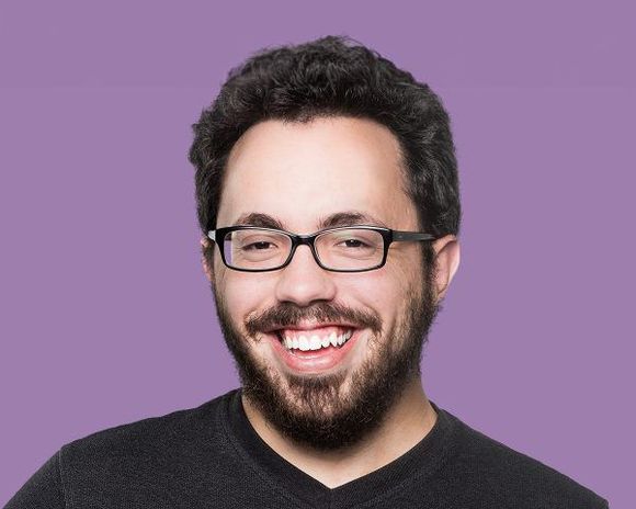 Man with glasses and beard smiles, purple background.