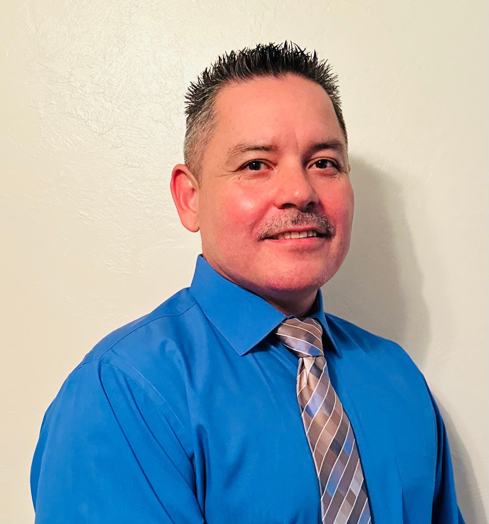 Man with dark hair and mustache wearing a blue shirt and patterned tie, smiling against a white background.