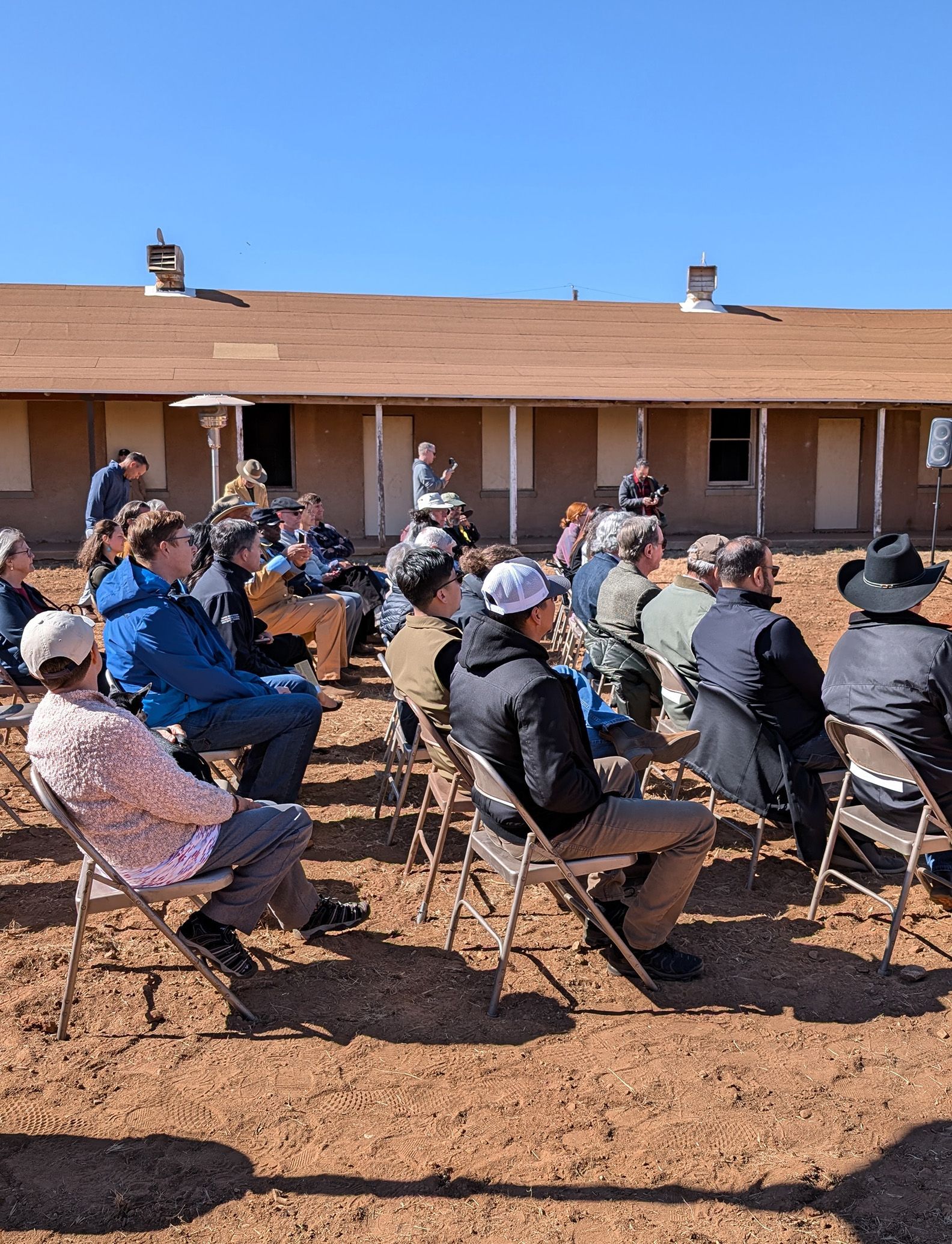 People seated outdoors in chairs, facing a building. Sunny day.