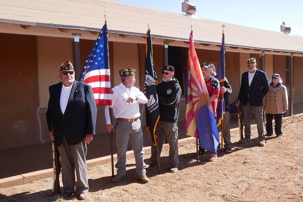 Veterans hold flags in front of a building. American flag and others are visible. Sunny day.
