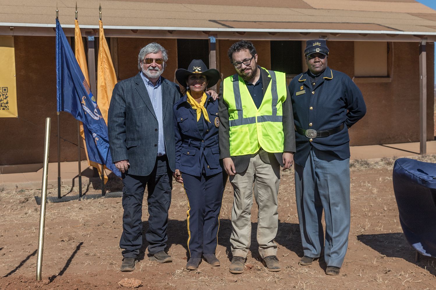 Four people stand outside a building; flags are behind them. Two wear uniforms.