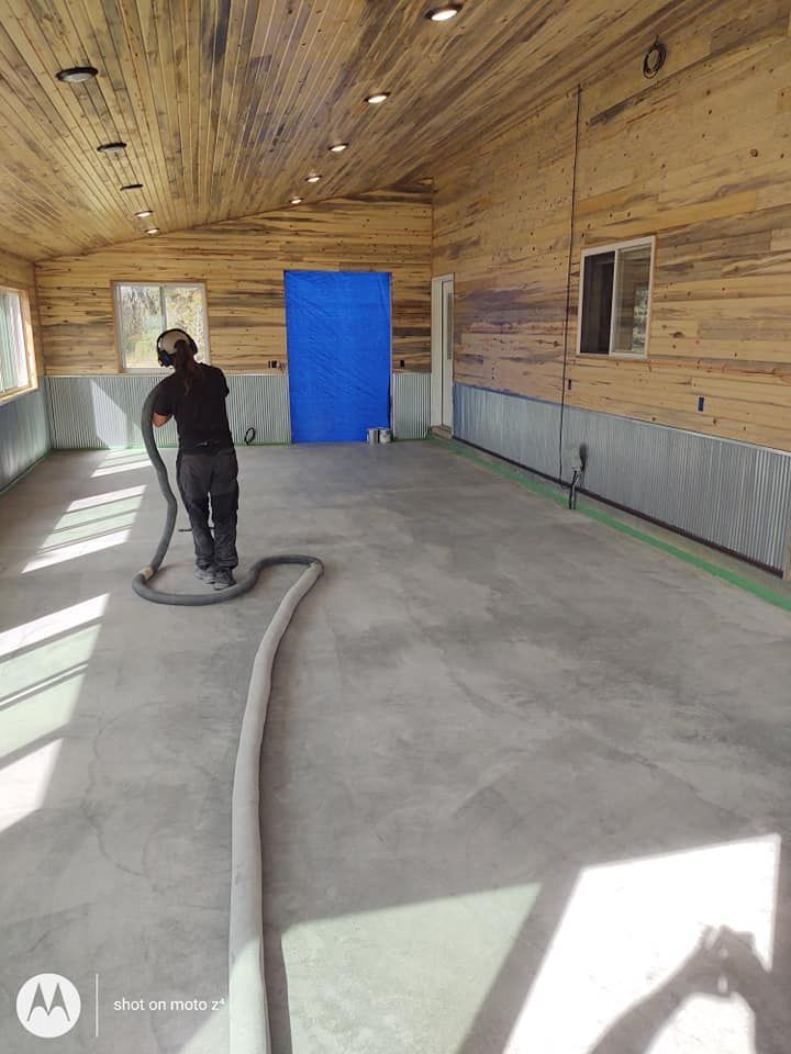 Worker smoothing a concrete floor inside a wood-paneled room with a blue door and windows.