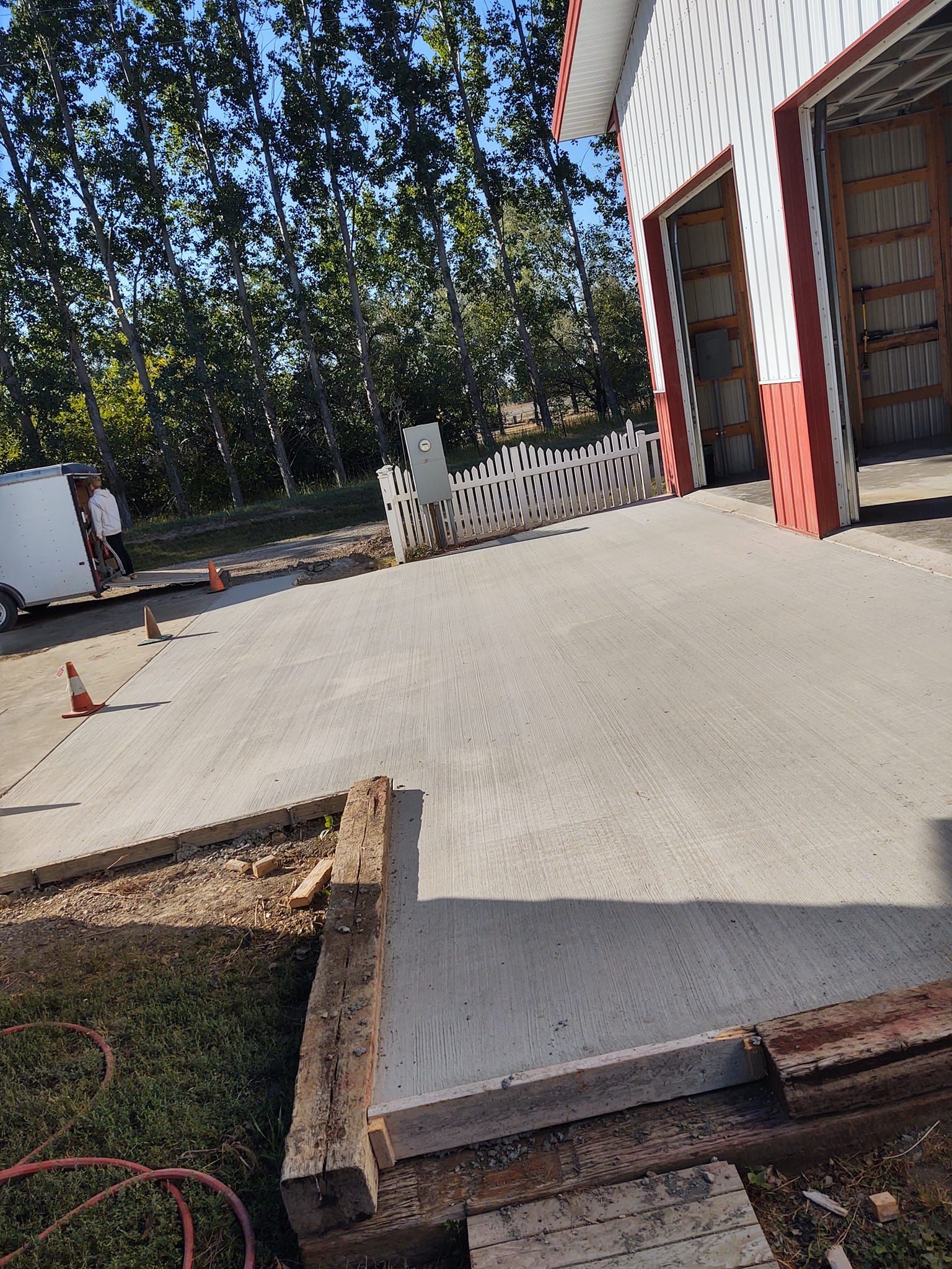 Concrete driveway leading to a red-trimmed garage, with a white fence and trees in the background.