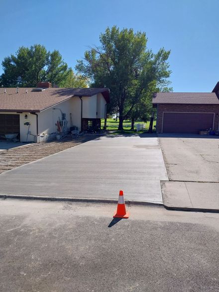 Driveway between two houses with an orange traffic cone in the foreground and trees in the background.