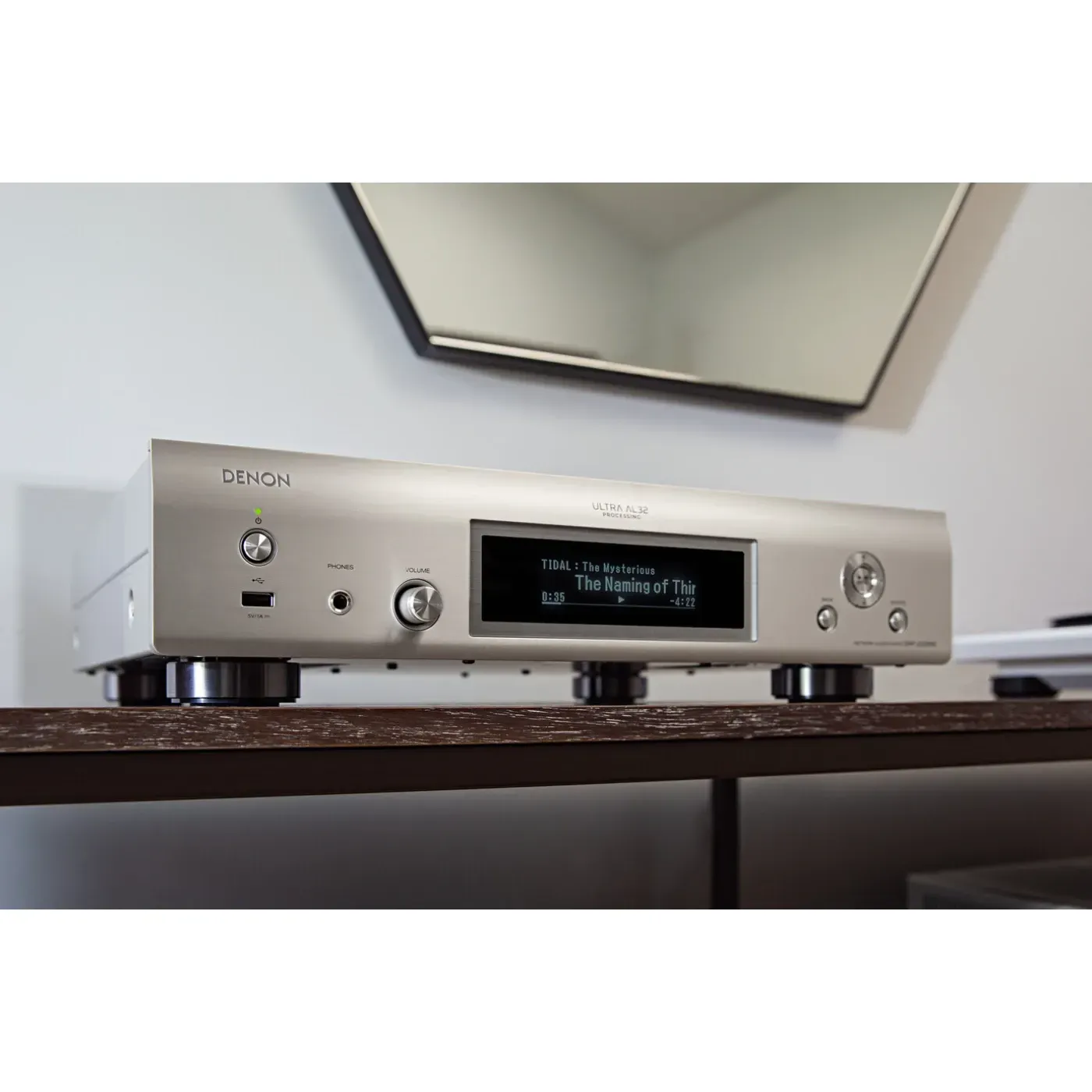 Silberne Audiokomponente mit beleuchtetem Display auf einem Holztisch unter einem Wandspiegel.