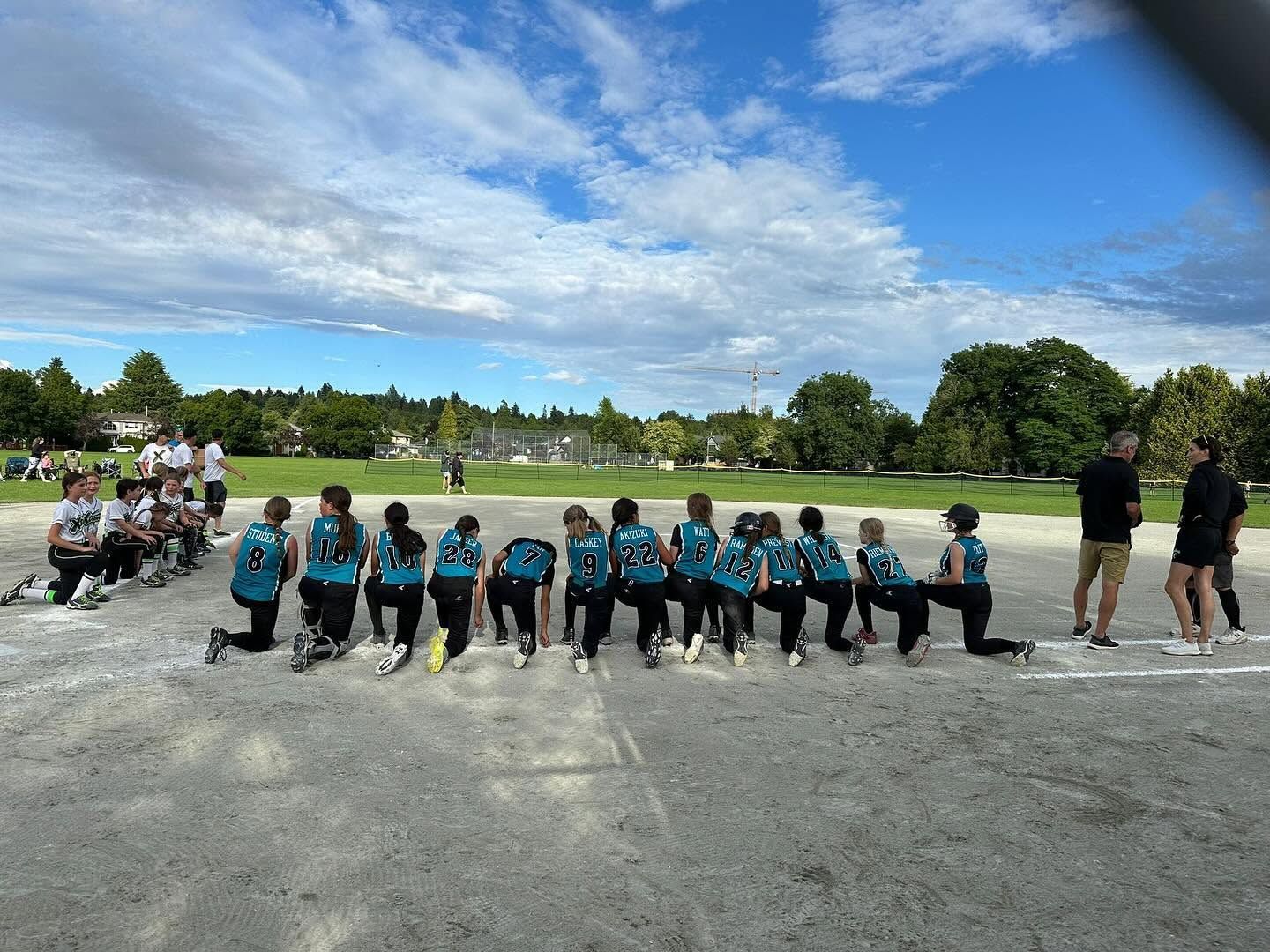 Children in teal shirts sit on benches at an outdoor sports field under a bright blue sky.