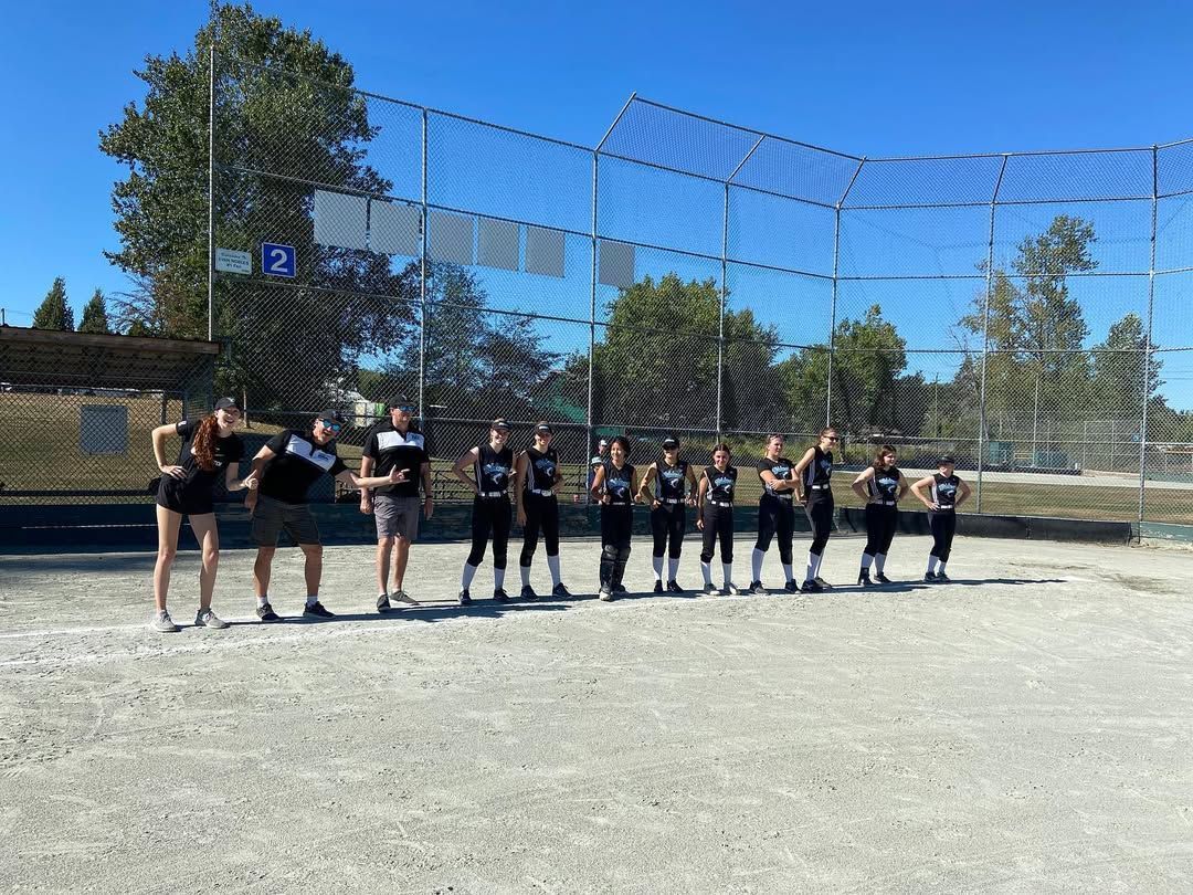 Group of people standing on a gravel field in front of a netted sports area under a clear blue sky.