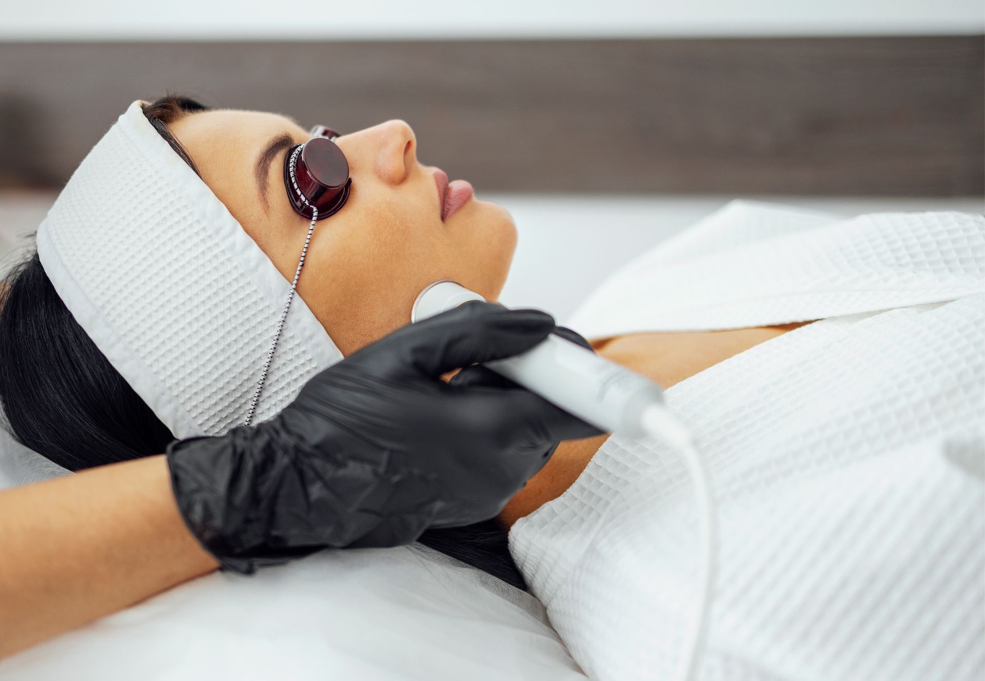 Mujer recibe tratamiento láser facial, usando gafas protectoras y diadema, en un entorno de spa.