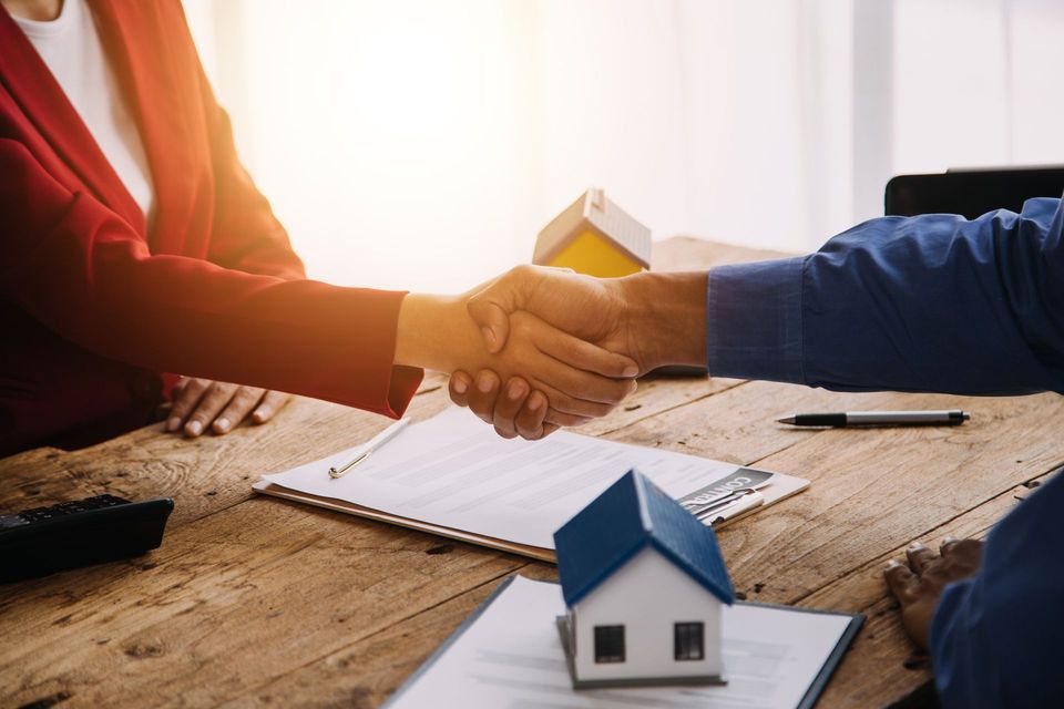 Two people shaking hands over a house model and paperwork on a wooden table, with a pen and notebook nearby