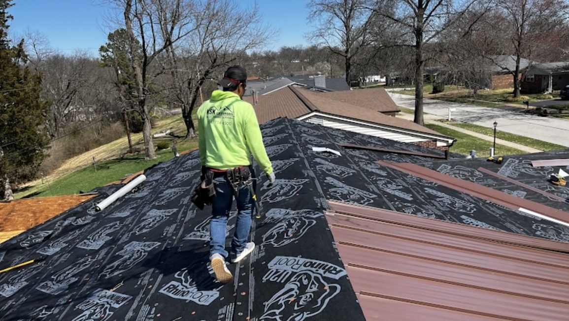 Worker in neon jacket walking on a roof under repair, with black underlayment and brown shingles nearby.