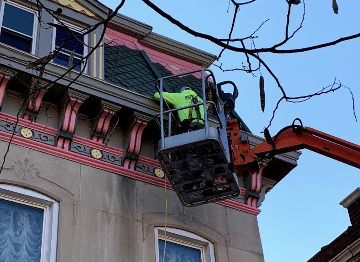 Worker in a lift basket repairs a building facade with an orange boom arm against a blue sky.