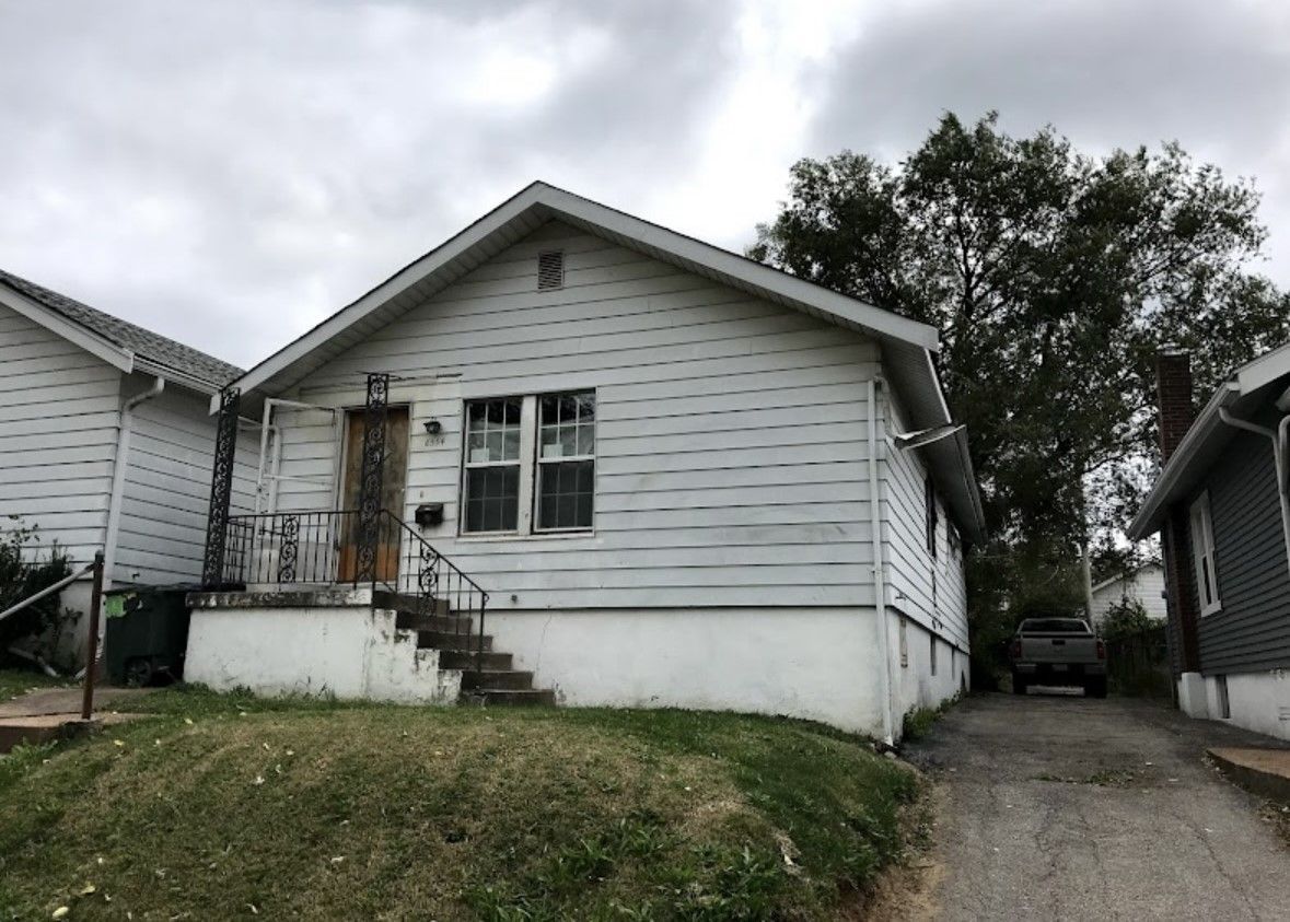 Small white house with front steps and overgrown yard beside a driveway, under cloudy skies