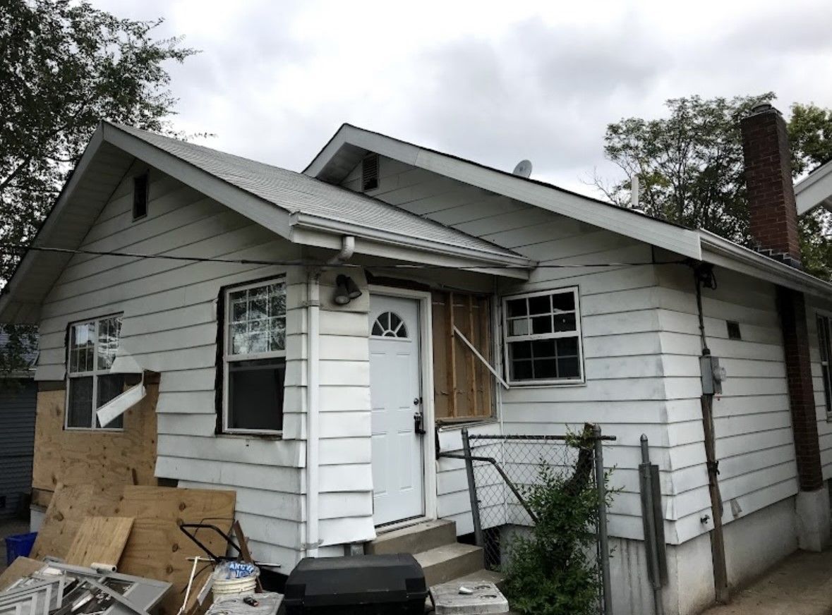 Small white house with damaged siding, boarded windows, and debris in the yard under a cloudy sky