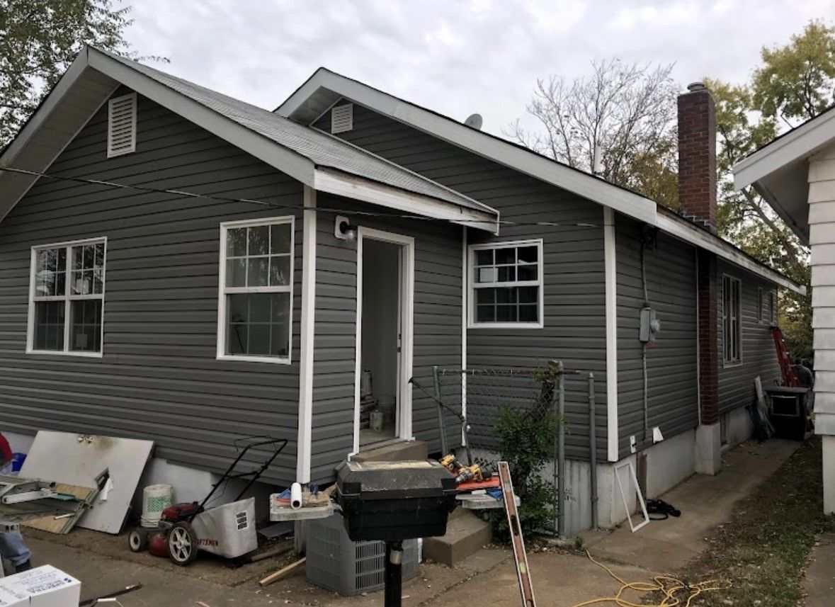 Gray house exterior with white trim, porch clutter, and a small fenced side yard