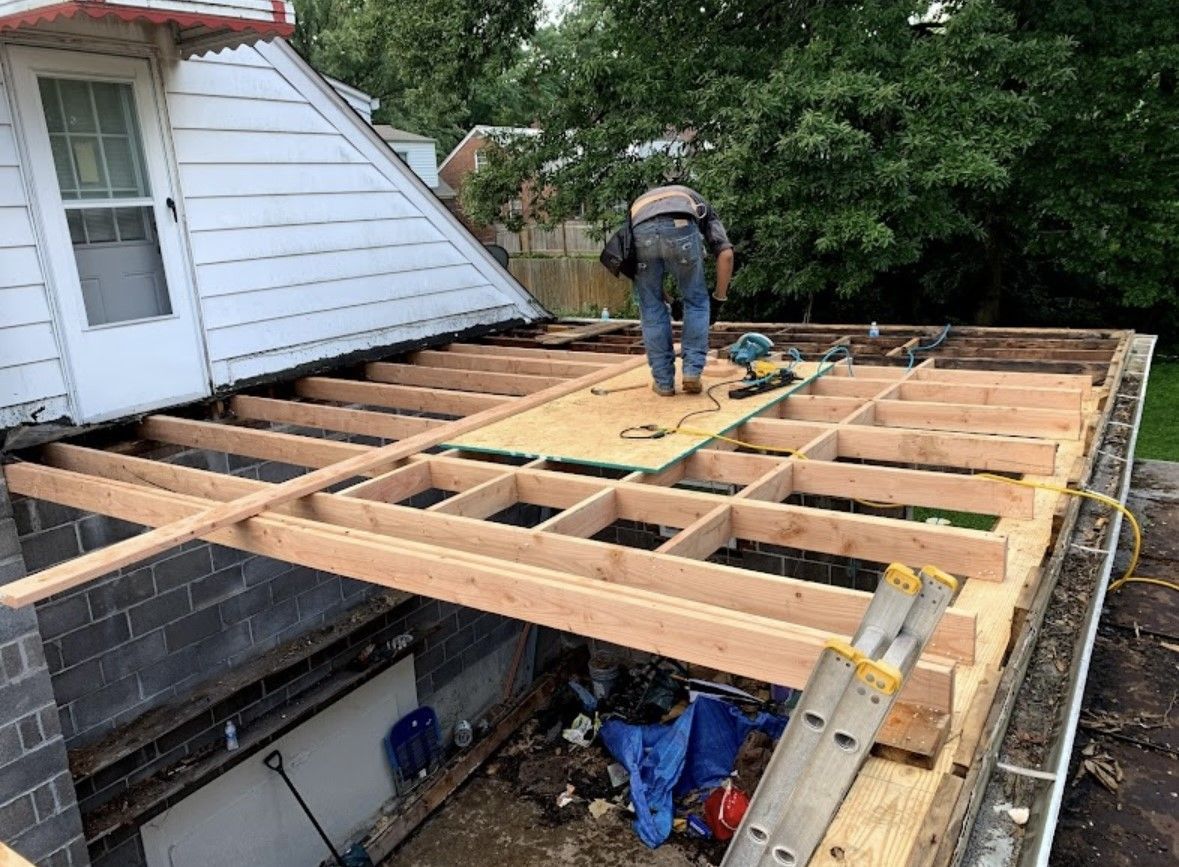 Worker framing a roof deck beside a house, with exposed wooden beams and tools on top.