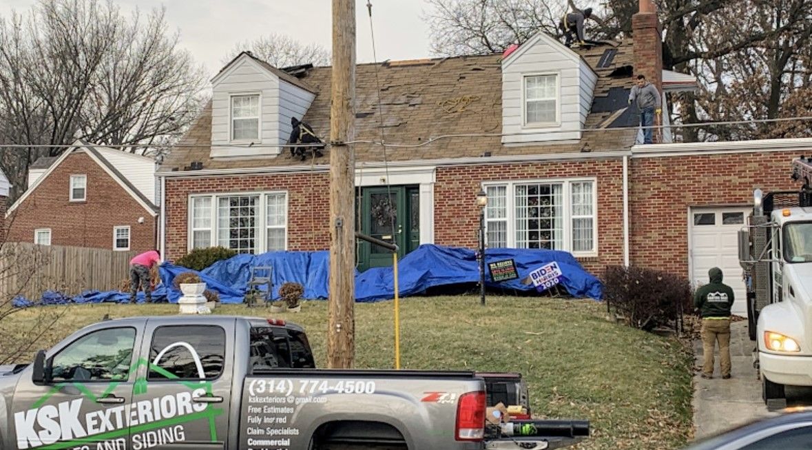 Brick house with blue tarps on the roof and a work truck parked in front, suggesting home repair.