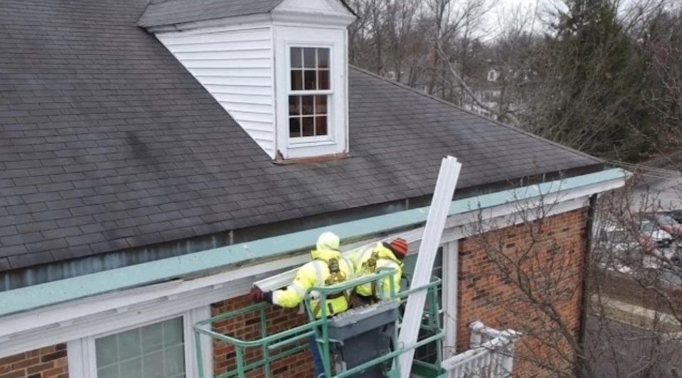 Workers in yellow safety gear on a roof beside a dormer window, using a lift near the gutters.