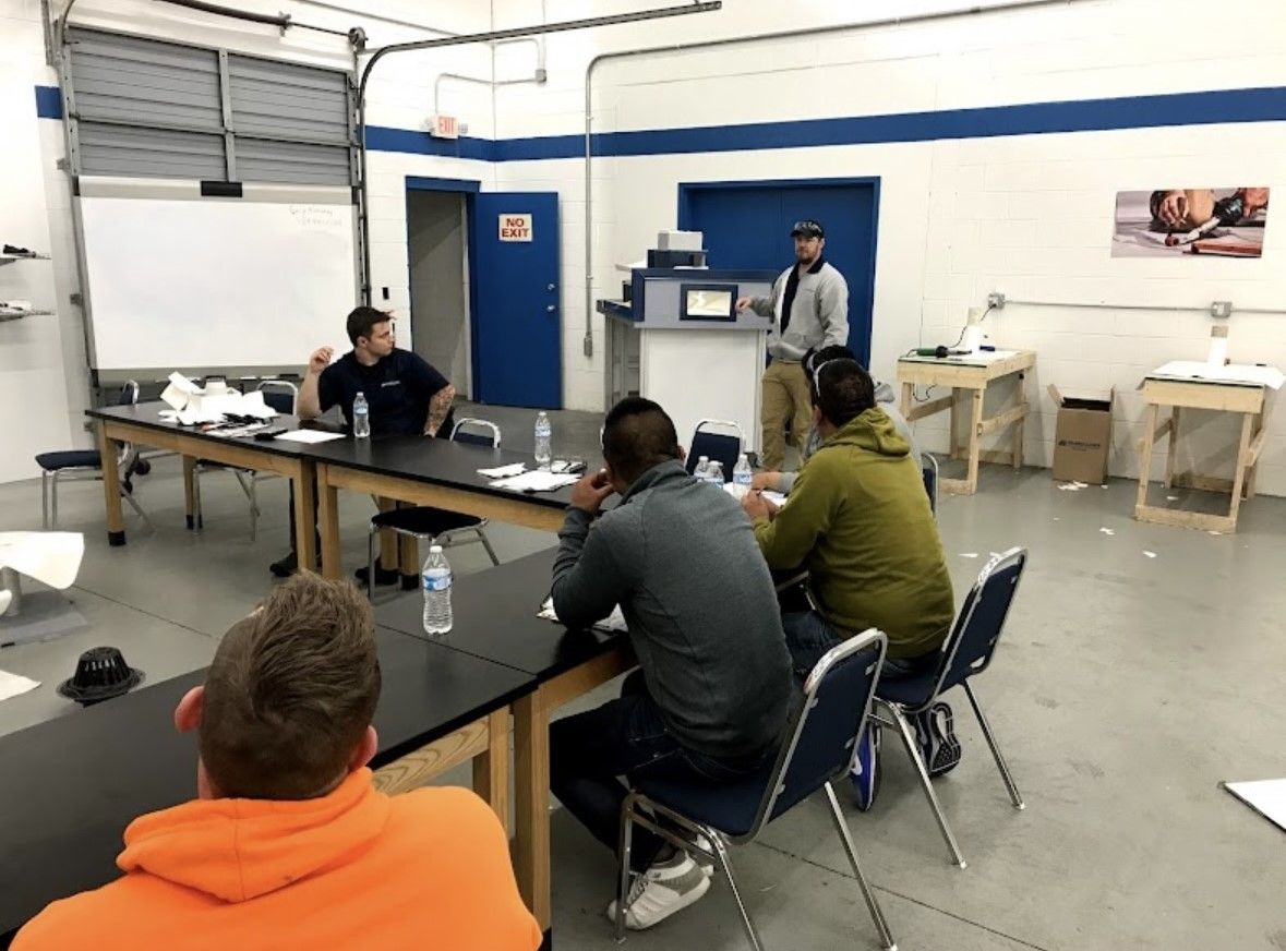 Small meeting in an industrial room with people seated at tables and a presenter near a whiteboard