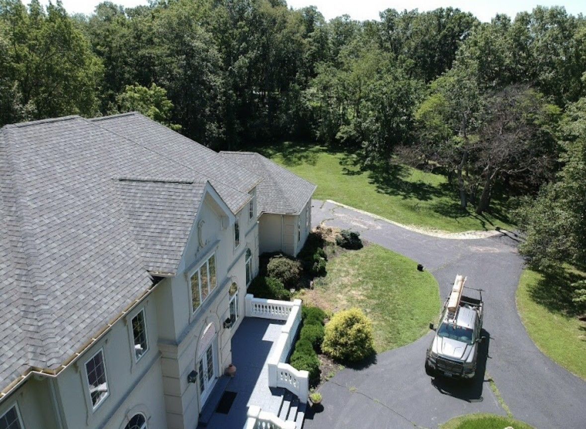 Aerial view of a large gray house with a driveway, patio, and wooded yard.