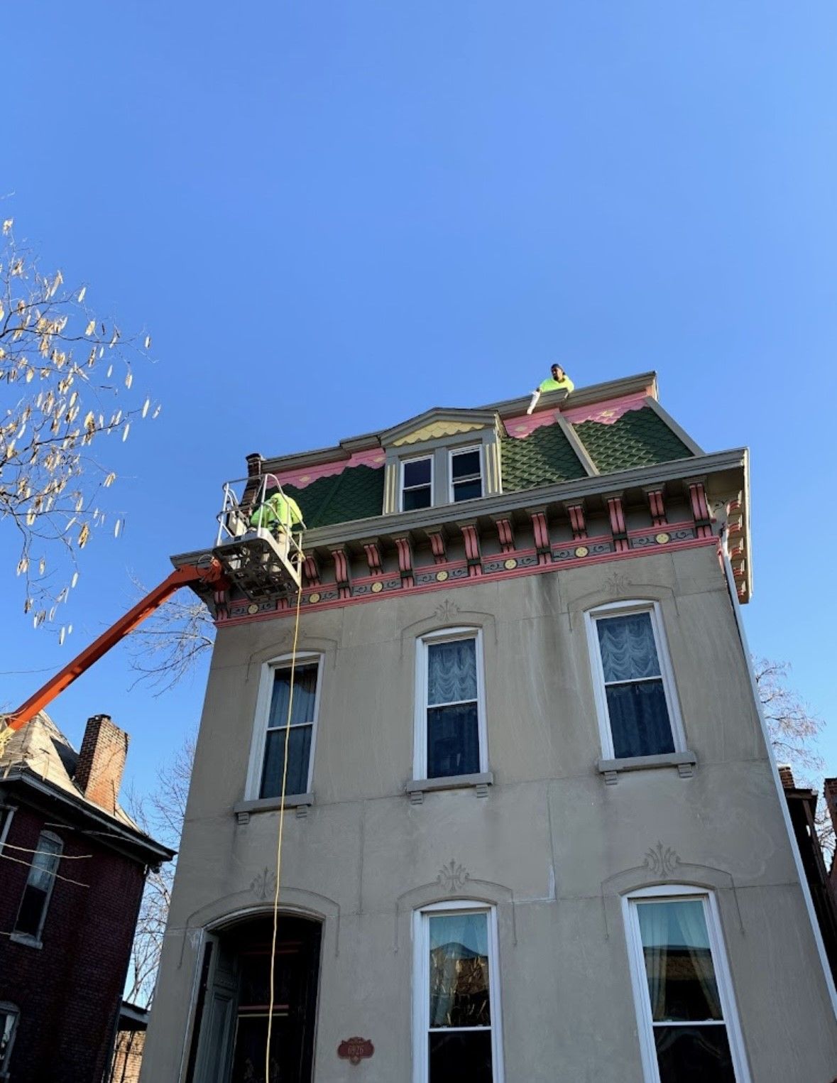Roofers working on a tall beige house with a green roof under a clear blue sky